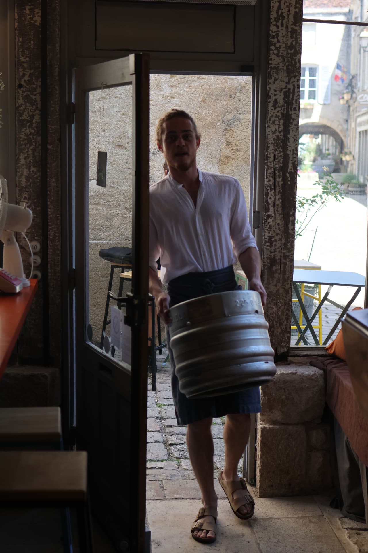 Un serveur rentre un fût de bière par la porte, la ruelle pavée en fond